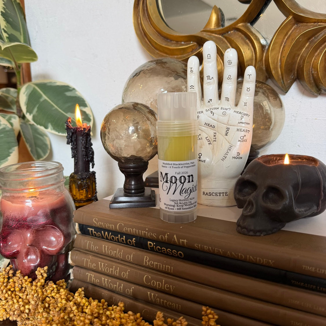 Moon Magick Legacy Menthol Salve in a wide styled scene with skull candle and vintage art books